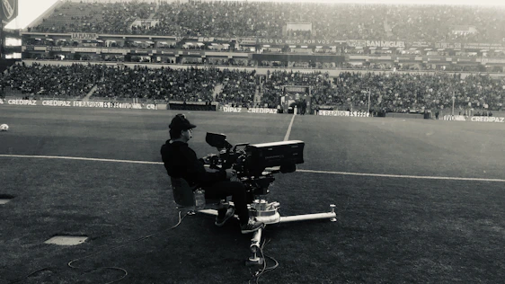 A dynamic black-and-white photo of a football creator filming content on the sidelines of a stadium at dusk.