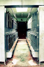 A narrow corridor lined with rows of metal lockers on both sides. The lockers are slightly worn, and the floor has visible signs of wear and discoloration. Overhead, fluorescent lighting partially illuminates the area, casting shadows in the dim space.