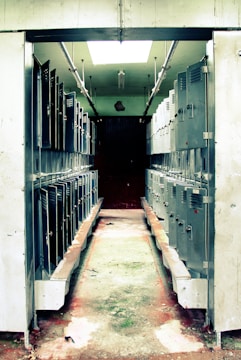 A narrow corridor lined with rows of metal lockers on both sides. The lockers are slightly worn, and the floor has visible signs of wear and discoloration. Overhead, fluorescent lighting partially illuminates the area, casting shadows in the dim space.