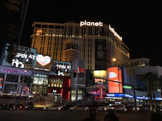 A nighttime scene of a bustling urban street featuring the brightly lit facade of the Planet Hollywood resort and casino. Neon lights and advertisements for shows and shops illuminate the area, with large digital screens displaying vibrant images and text. Cars and people are visible in the foreground, adding to the lively atmosphere.