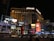 A nighttime scene of a bustling urban street featuring the brightly lit facade of the Planet Hollywood resort and casino. Neon lights and advertisements for shows and shops illuminate the area, with large digital screens displaying vibrant images and text. Cars and people are visible in the foreground, adding to the lively atmosphere.