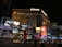 A nighttime scene of a bustling urban street featuring the brightly lit facade of the Planet Hollywood resort and casino. Neon lights and advertisements for shows and shops illuminate the area, with large digital screens displaying vibrant images and text. Cars and people are visible in the foreground, adding to the lively atmosphere.