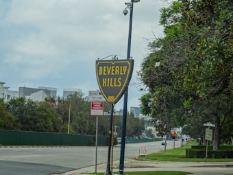 A Beverly Hills city limit sign is prominently displayed on a street with trees and bushes lining the sidewalks. In the background are some buildings and a cloudy sky.