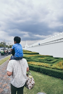 A child sits on the shoulders of an adult walking along a landscaped path next to a tall, white decorative wall. The sky is overcast with thick, gray clouds, suggesting a cloudy day. The adult carries a polka-dotted bag while the well-groomed hedges run parallel to the path. Tuk-tuk vehicles are visible on the street nearby.