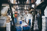 A bustling indoor market scene with numerous white styrofoam boxes stacked along the narrow aisle. Several ladders and hanging cables create a busy and industrious atmosphere. A blue plastic crate filled with red fish or seafood is visible in the foreground. The lighting is dim, suggesting an early morning market setting.