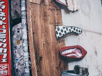 The image showcases a section of a textured wall with multiple shoe designs displayed vertically. One shoe has a black and white polka dot pattern, another is red with gold studs, and a third is gray with a fur detail. The left side features a pole covered in various stickers and a vertical red sign with Japanese characters.