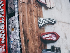 The image showcases a section of a textured wall with multiple shoe designs displayed vertically. One shoe has a black and white polka dot pattern, another is red with gold studs, and a third is gray with a fur detail. The left side features a pole covered in various stickers and a vertical red sign with Japanese characters.