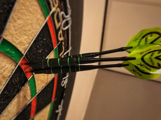 Three darts are embedded in a dartboard, each landing in the red inner ring. The dartboard features multiple colors, including black, green, and red sections, with visible wire dividers. The darts have yellow flights with brand markings.