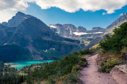 A scenic mountain landscape featuring a rugged trail leading through lush greenery towards a turquoise lake surrounded by towering peaks. The mountain range is capped with snow, and the sky is dotted with fluffy clouds.