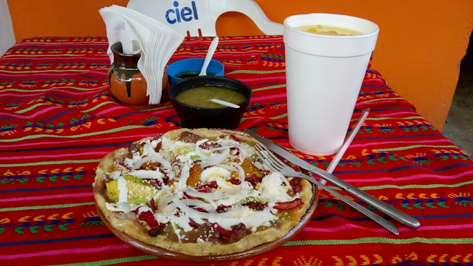 A colorful tablecloth with a traditional design covers a table set with a large, white takeout cup, a ceramic vase holding napkins, a small black bowl of green sauce, and a plate with a Mexican dish topped with cheese, onions, and avocado slices.