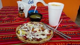 A colorful tablecloth with a traditional design covers a table set with a large, white takeout cup, a ceramic vase holding napkins, a small black bowl of green sauce, and a plate with a Mexican dish topped with cheese, onions, and avocado slices.