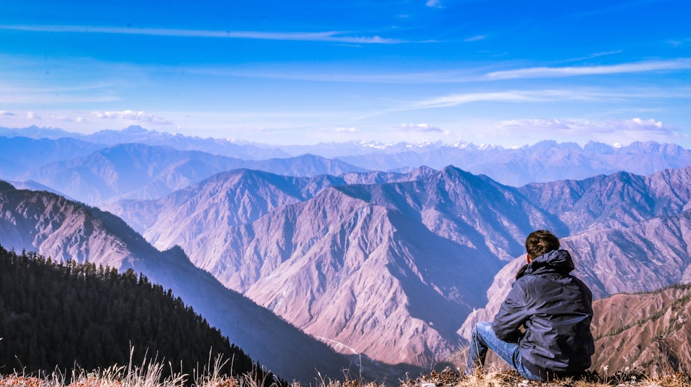 man sitting at the edge of a mountain facing the mountains during day
