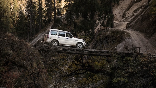 A white SUV crosses a narrow wooden bridge in a rugged mountain landscape, surrounded by dense forest and rough terrain. The scene captures a sense of adventure and exploration in a remote and challenging environment.