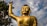 A large golden statue of Buddha with its right arm raised stands prominently against a backdrop of a cloudy blue sky and green foliage.