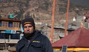 A man wearing a dark jacket with 'NEPAL POLICE' written on it is standing outdoors. He has a black cap with the word 'COMMANDO' and a small red mark on his forehead. The background consists of several buildings, a red tent-like structure, and hills with sparse trees.