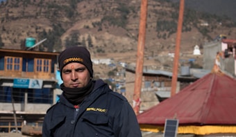 A man wearing a dark jacket with 'NEPAL POLICE' written on it is standing outdoors. He has a black cap with the word 'COMMANDO' and a small red mark on his forehead. The background consists of several buildings, a red tent-like structure, and hills with sparse trees.