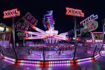 A vibrant amusement park ride featuring bright, colorful lights arranged in patterns. Several mechanical arms extend outward with seats attached, and signs with 'XXL' are prominently displayed. The scene is set against a dark nighttime backdrop, amplifying the vividness of the neon lights.
