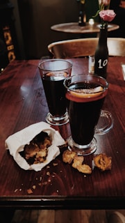 Two glasses filled with a dark beverage, likely mulled wine, are placed on a wooden table with orange slices in them. A paper bag spills out roasted chestnuts beside the glasses. A black bottle with a pink flower and the number 10 sits in the background, enhancing the cozy and intimate atmosphere of the setting.
