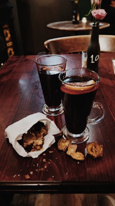 Two glasses filled with a dark beverage, likely mulled wine, are placed on a wooden table with orange slices in them. A paper bag spills out roasted chestnuts beside the glasses. A black bottle with a pink flower and the number 10 sits in the background, enhancing the cozy and intimate atmosphere of the setting.