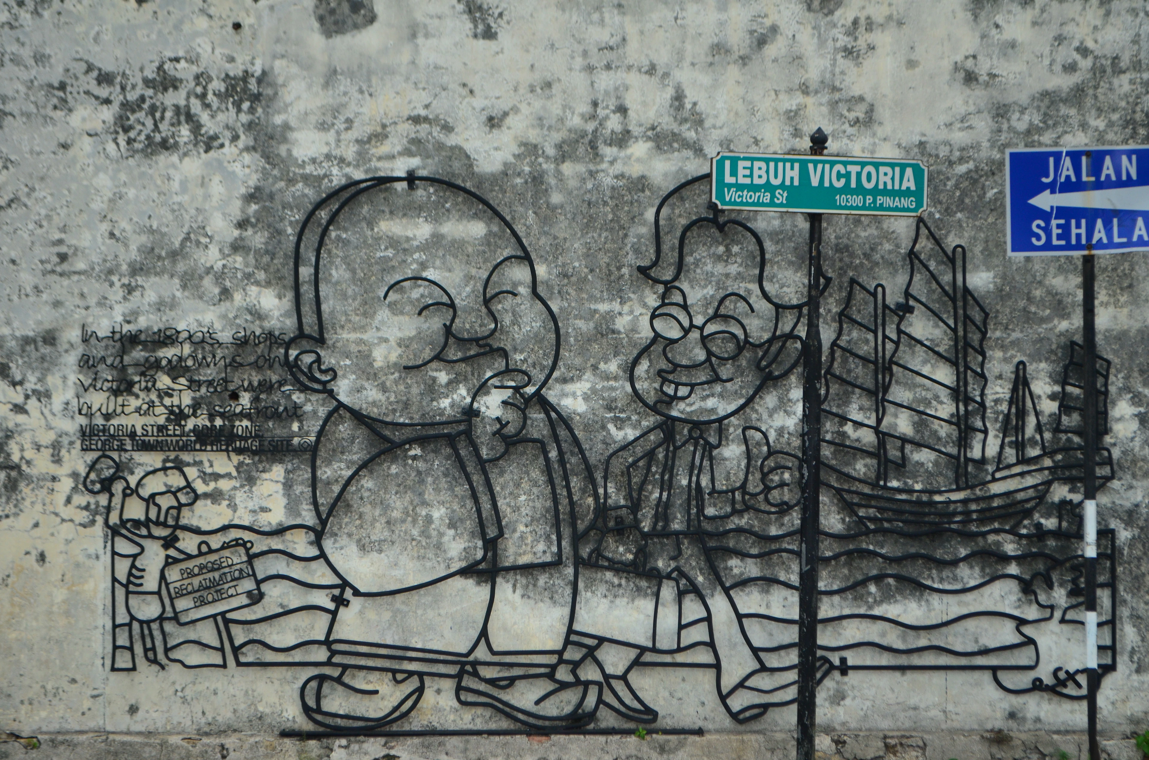 A steel-rod caricature street artwork of two figures and a Chinese junk, mounted on a weathered wall beneath the Lebuh Victoria street sign in George Town, Penang