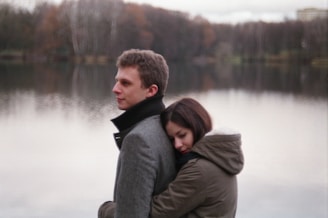 A young couple stands by a serene lake with forested background. The woman is hugging the man from behind, resting her head on his back. Both are dressed in warm coats, suggesting a chilly day.
