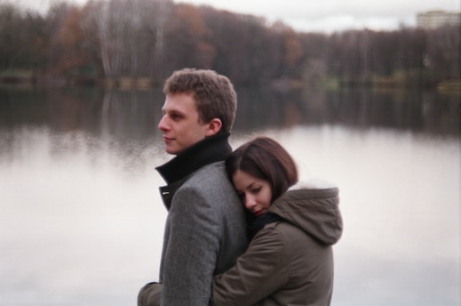 A young couple stands by a serene lake with forested background. The woman is hugging the man from behind, resting her head on his back. Both are dressed in warm coats, suggesting a chilly day.