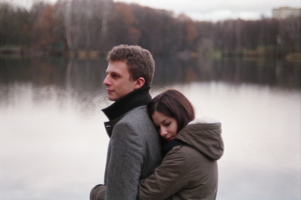 woman hugging man near body of water and trees during daytime