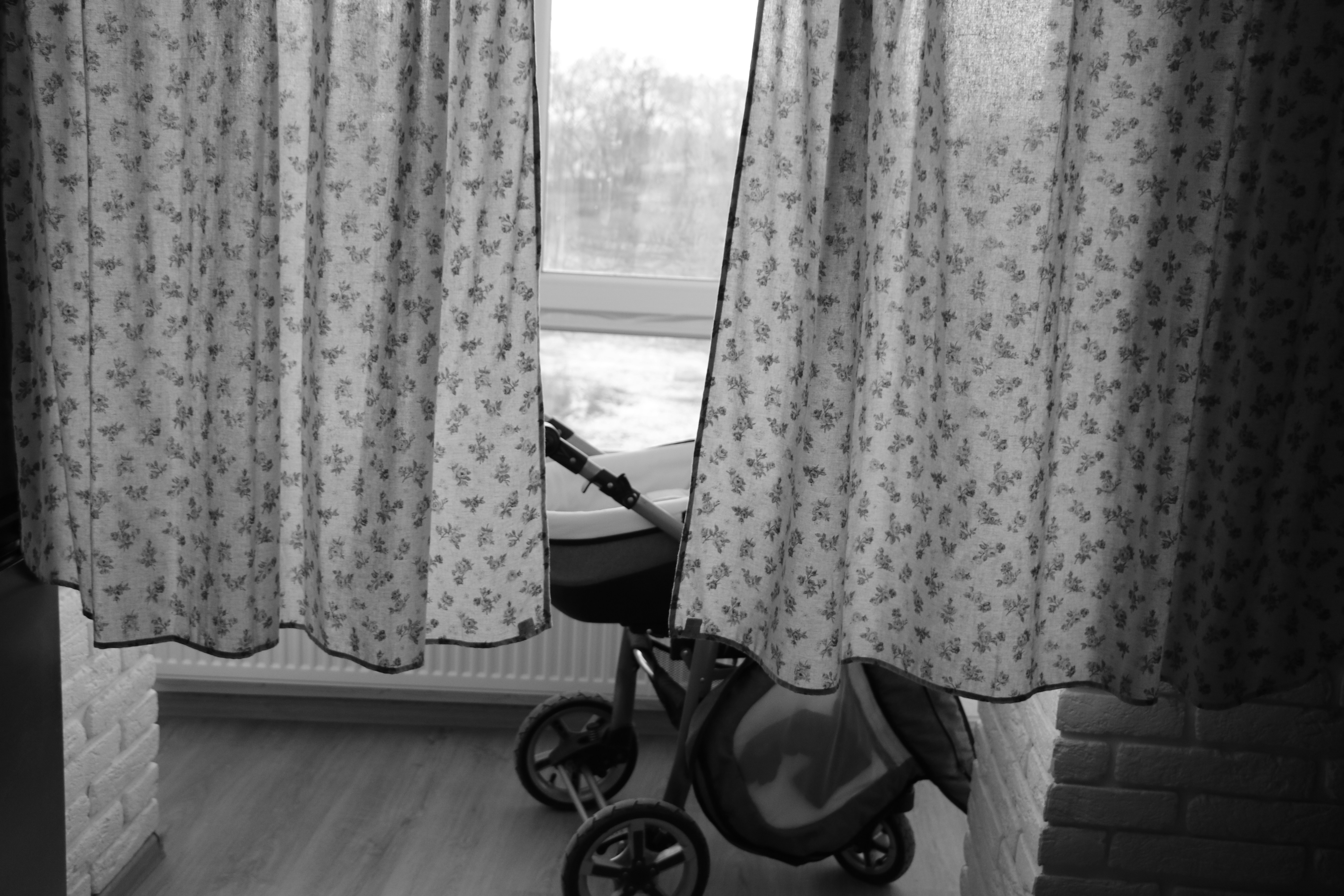 A dimly lit room features floral-patterned curtains partially drawn open, revealing a view of a window. Behind the curtains, a baby stroller is visible, placed on a wooden floor next to a white brick wall.