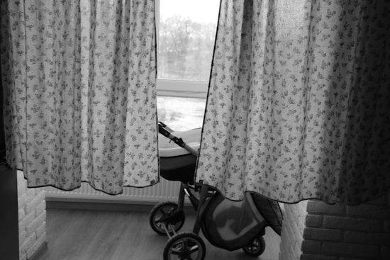 A dimly lit room features floral-patterned curtains partially drawn open, revealing a view of a window. Behind the curtains, a baby stroller is visible, placed on a wooden floor next to a white brick wall.