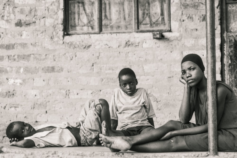 grayscale photo of three person sitting and standing in front of concrete wall