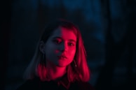 A person with shoulder-length hair is illuminated by a deep red light against a dark blue background. The contrast between the red and blue tones creates a striking visual effect.