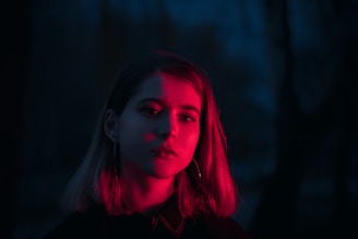 A person with shoulder-length hair is illuminated by a deep red light against a dark blue background. The contrast between the red and blue tones creates a striking visual effect.