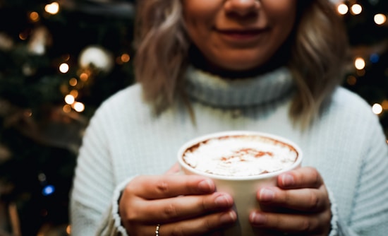 A person wearing a cozy sweater holds a cup of frothy hot chocolate or coffee with both hands. The background features blurred glowing lights, suggesting a festive or warm setting.