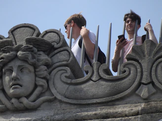 A detailed stone sculpture with a face and intricate patterns is in the foreground. Behind the sculpture, two individuals stand holding onto a metal barrier—one is gazing into the distance while the other looks at a smartphone. The scene takes place outdoors on a sunny day.