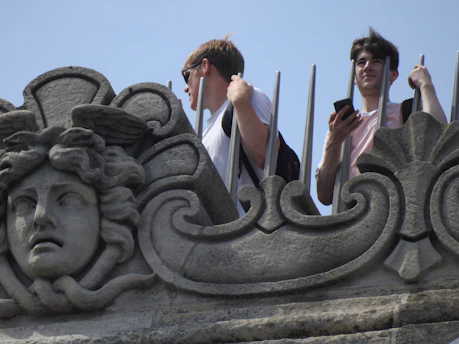 A detailed stone sculpture with a face and intricate patterns is in the foreground. Behind the sculpture, two individuals stand holding onto a metal barrier—one is gazing into the distance while the other looks at a smartphone. The scene takes place outdoors on a sunny day.