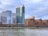 A waterfront view of modern high-rise buildings including a tall structure with a distinctive red letter 'W' at the top, surrounded by other residential and office buildings. The scene is calm with a partly cloudy sky and a gently flowing river in the foreground.