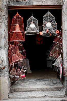 A collection of colorful nets are displayed hanging around an open doorway. The nets have various shapes and contain objects like floats inside them, creating an interesting pattern. The entrance is framed by rough, weathered stone, adding a rustic charm.