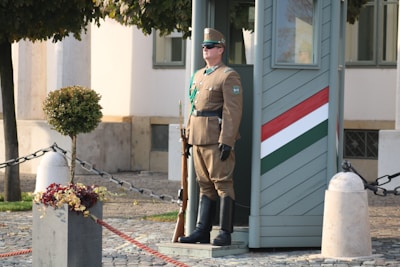 A uniformed guard stands solemnly beside a small sentry box painted with a tricolor stripe, likely representing a national flag. The scene is set outdoors with a cobblestone ground, decorative plant urns, and some greenery. The guard holds a rifle and is wearing sunglasses and a military hat.