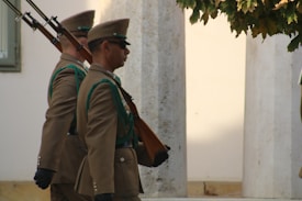 Two uniformed guards are marching in a solemn and synchronized manner, holding rifles upright on their shoulders. They are wearing brown military attire with green accents and black gloves. The background includes stone pillars and some green foliage, suggesting an outdoor setting near a formal or ceremonial location.