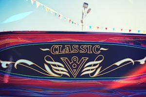 A vibrant design labeled 'Classic' with ornate golden script and a stylized 'V8' emblem on a deep navy background. Surrounding this is a swirl of artistic patterns in red and blue. Above, a string of colorful triangular flags points towards the clear sky, and a lamp post is visible.