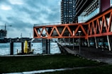 An urban waterfront scene featuring modern architecture with bright orange geometric structures extending over a walkway. The background shows a body of water with industrial elements, including a construction crane and a tall building. The scene conveys a blend of nature and urban development.