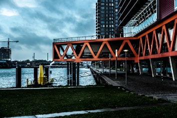 An urban waterfront scene featuring modern architecture with bright orange geometric structures extending over a walkway. The background shows a body of water with industrial elements, including a construction crane and a tall building. The scene conveys a blend of nature and urban development.