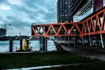 An urban waterfront scene featuring modern architecture with bright orange geometric structures extending over a walkway. The background shows a body of water with industrial elements, including a construction crane and a tall building. The scene conveys a blend of nature and urban development.