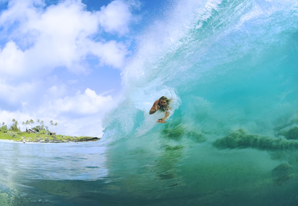 A surfer rides inside the curve of a translucent turquoise wave, creating a dynamic and thrilling scene. The bright blue sky and fluffy white clouds add a sense of openness and adventure, while palm trees and a rocky shoreline are visible in the distance, suggesting a tropical location.
