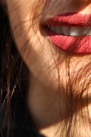 A close-up of lips with natural makeup, slightly parted, showing teeth. Hair strands cross over the face, partially obscuring the view and creating a soft, intimate feel. The lighting casts gentle shadows across the skin, highlighting the texture of the lips.