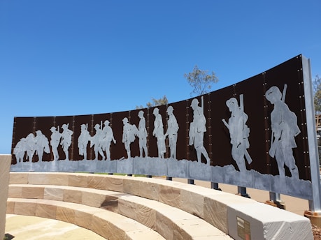 A public art installation featuring a series of metallic silhouettes depicting soldiers standing in various poses. These figures are mounted on a curved panel and arranged in a procession against a backdrop of clear blue sky. The installation is situated in a landscaped area with circular stone seating in the foreground.