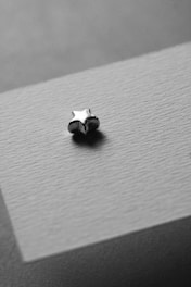 A small, metallic star-shaped object rests on a textured surface in grayscale. The lighting creates a soft shadow, emphasizing the object's smooth and reflective properties.