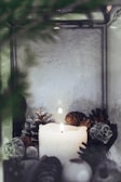 A white candle is situated at the center, glowing softly amidst an assortment of decorative pinecones and twigs. The setting appears to be tranquil, with muted colors and a blurred background. The elements are arranged in a rustic, cozy manner inside a metallic frame.