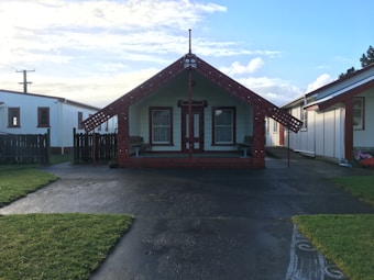 A traditional Maori meeting house is centrally located, showcasing intricate carvings and designs on its wooden structure. It is surrounded by grass, with paved pathways leading up to the entrance. Nearby buildings have white exteriors with similar architectural features, and there's a clear blue sky with a few clouds.