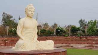 A large, serene statue of Buddha made from a light-colored stone sits on a circular platform in an open area. The statue is surrounded by greenery and trees, with an amphitheater-style seating structure made from red stone in the background. The environment is peaceful, with overcast skies above.